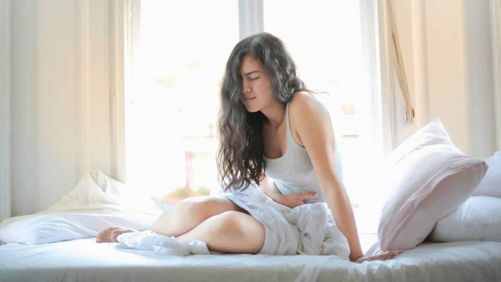 A woman with long dark hair is sitting on a bed in a bright room, clutching her abdomen with a pained expression. She is dressed in a gray tank top and surrounded by white bedding and pillows. Sunlight streams through the large windows in the background.