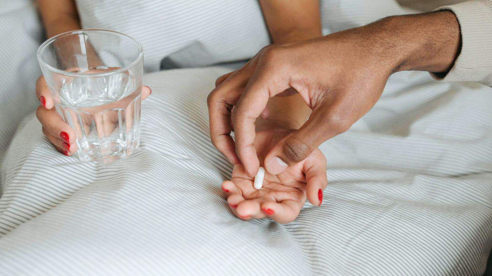 Close-up of a person lying in bed holding a glass of water in their right hand, while another hand places a white pill in the person's left. The background shows white-striped bedding.