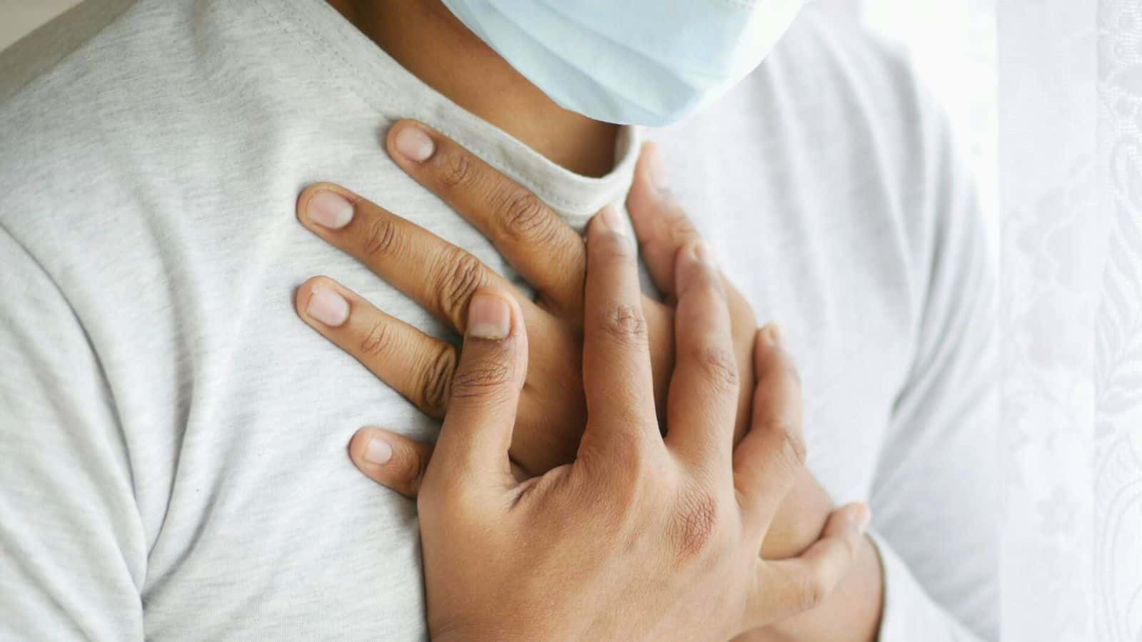 A person wearing a light grey shirt and a blue face mask is pictured. They have both hands placed on their chest, overlapping each other. The background appears to be softly lit and partially covered by a sheer white curtain.