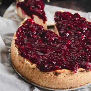 A cheesecake topped with a thick layer of cherry compote sitting on a piece of parchment paper. A slice has been cut and removed, revealing the creamy texture inside. The edges of the cheesecake have a light brown crust.