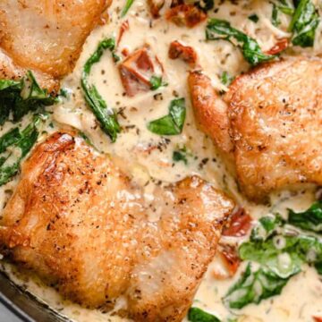 A close-up of a creamy dinner recipe features golden-brown chicken thighs, fresh spinach, and diced sun-dried tomatoes in a white sauce. This meal is cooked in a skillet, displaying a rich and savory presentation that's perfect for curing takeout addictions.