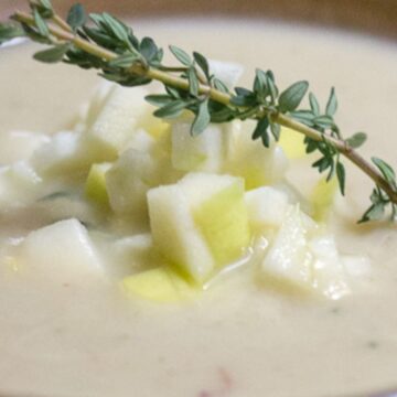 A close-up of a bowl of creamy soup garnished with diced green apples and a sprig of fresh thyme. This Retro One-Pot Classic, with its smooth, light-colored base, likely features a blend of vegetables or potatoes.