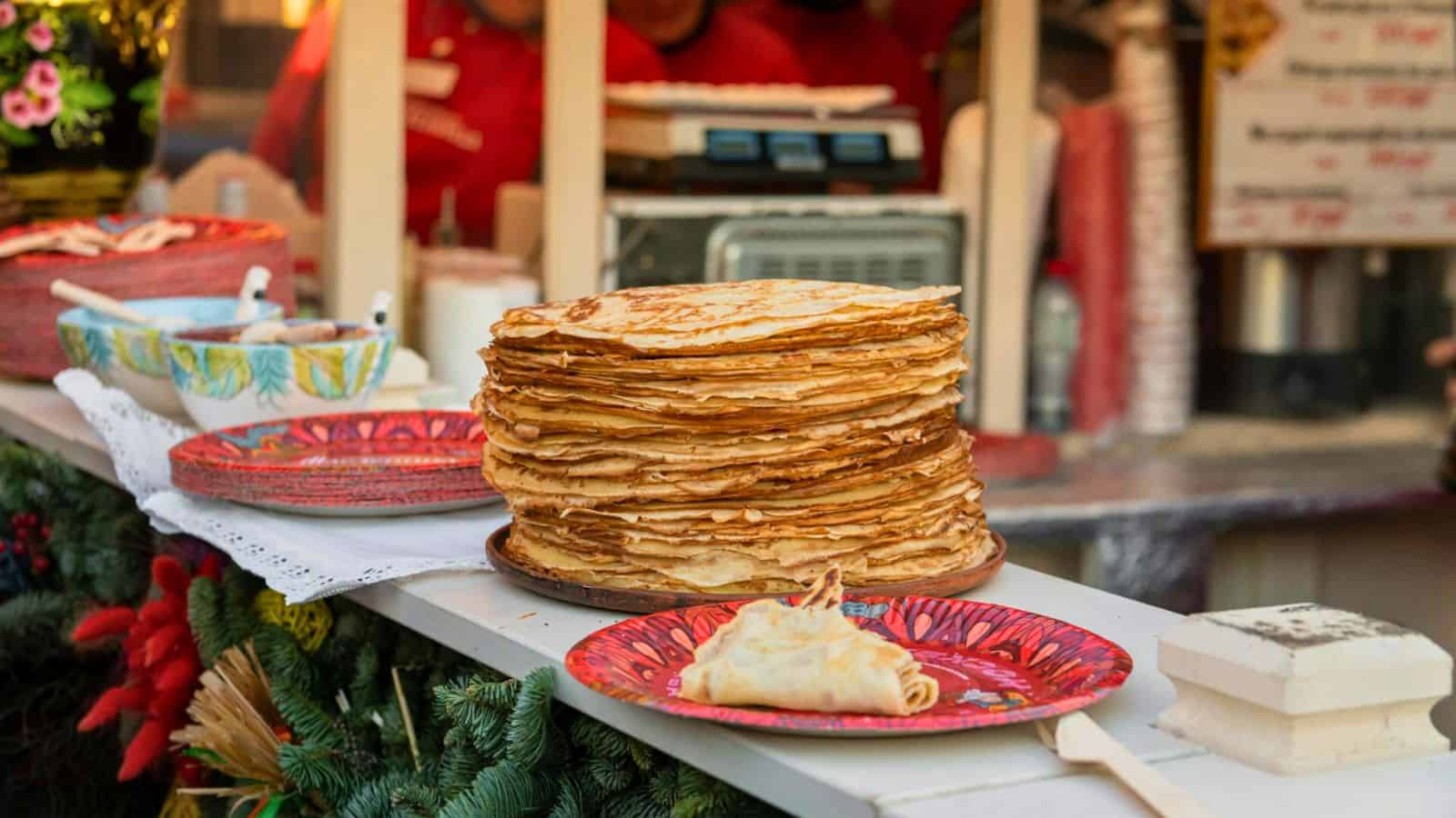 A tall stack of crepes is displayed on a counter decorated with Christmas-themed ornaments and greenery. Nearby, a plate holds some rolled crepes. In the background, blurred figures and a menu board.