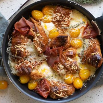 A skillet contains a baked dish with croissants, yellow cherry tomatoes, sliced ham, melted cheese, and sprigs of herbs, resting on a gray cloth. This delightful creation is inspired by classic casserole recipes. The surface underneath is light-colored with scattered grains.
