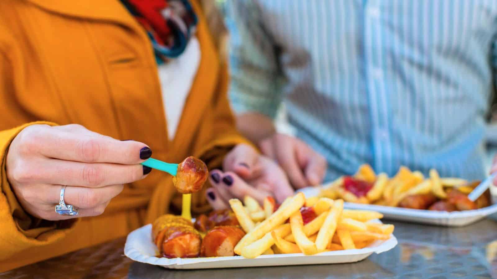 Two people enjoy a meal at a table. One holds a fork with a piece of sausage, and in front of them, there's a tray of iconic street foods like sausages and fries. The other has their tray topped with ketchup-covered sausages and fries. Only their torsos and arms are visible in this snapshot.