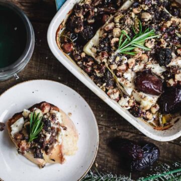 A casserole dish filled with a baked cheese and nut dish garnished with rosemary sprigs sits on a wooden table beside a small plate with a single serving of the dish. A green candle and pine branches are nearby, adding to a rustic table setting.