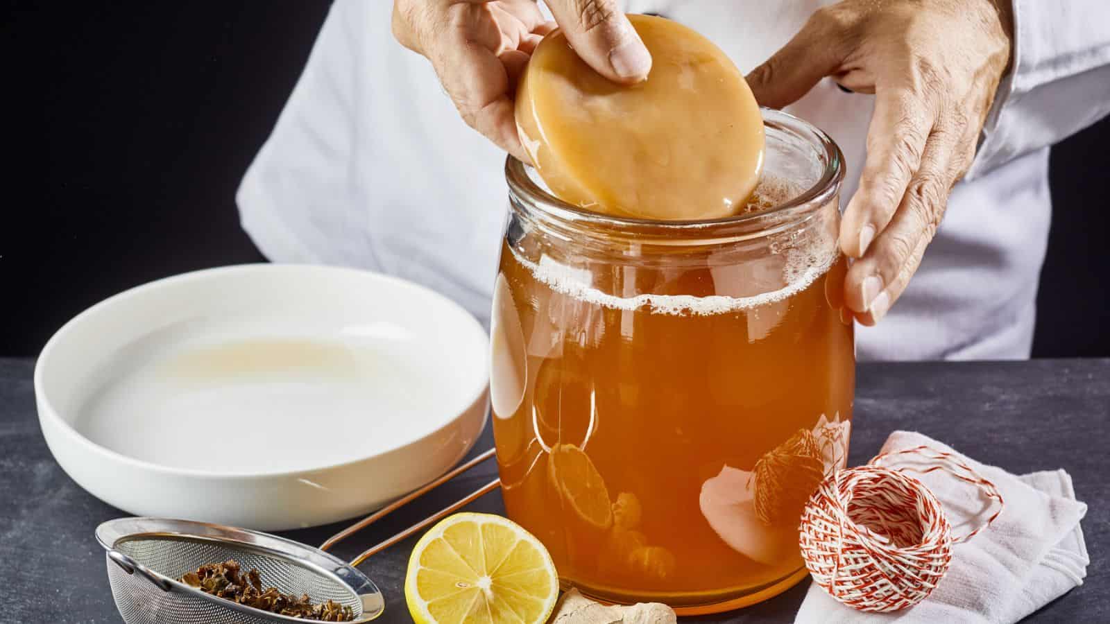 A person is placing a kombucha SCOBY into a large glass jar filled with kombucha tea. A white bowl, lemon half, string, and tea strainer with herbs are on the side. The background is dark and the person is wearing a white garment.