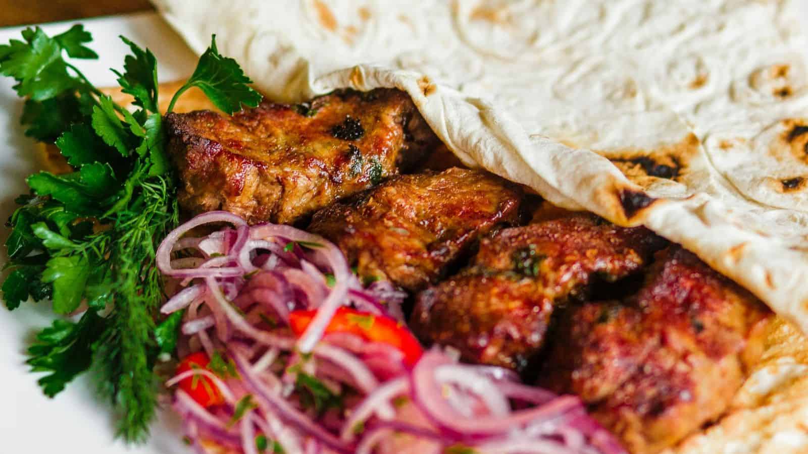 A close-up shot of a meal featuring grilled pieces of meat partially wrapped in flatbread. The dish is garnished with fresh parsley and dill, accompanied by thinly sliced red onions and bell peppers. A traveler’s delight served on a white plate.