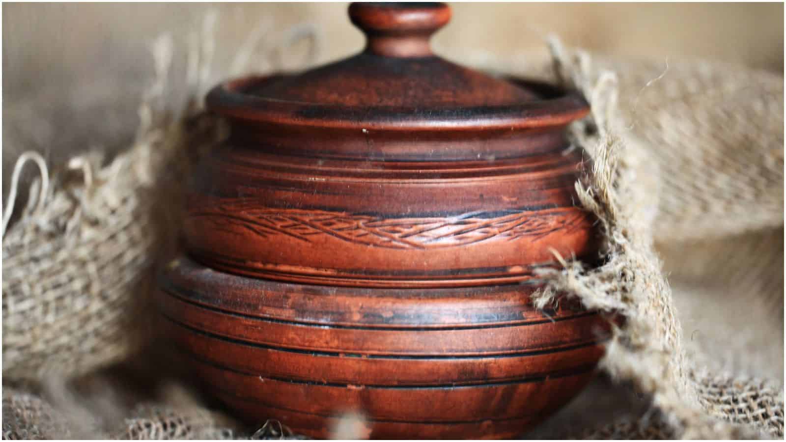 A close-up of a rustic, brown clay pot with intricate carvings on its surface. The pot has a lid and is surrounded by coarse burlap fabric.