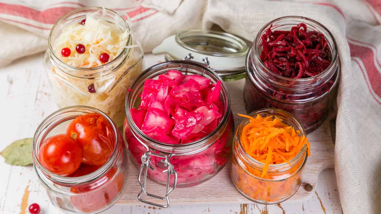 An assortment of pickled vegetables is displayed in glass jars on a white wooden surface. The jars contain pickled cherry tomatoes, shredded carrots, shredded beets, cabbage with cranberries, and pink-colored cabbage. A cloth is partially visible in the background.