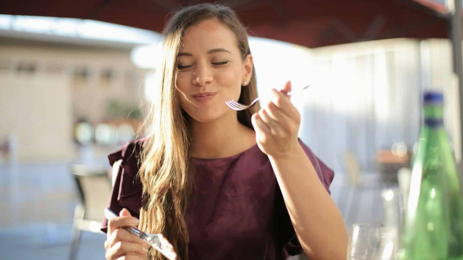 A woman with long hair is sitting outdoors under an umbrella, wearing a purple blouse. With her eyes closed and a fork close to her mouth. A bottle and a glass are on the table in front of her.