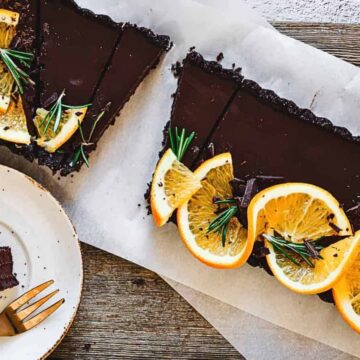 A chocolate tart topped with orange slices and rosemary sprigs is partially sliced on a wooden table, embodying the essence of summer treats. Nearby, a small plate holds a slice of the tart with a golden fork, while the dessert rests elegantly on parchment paper.