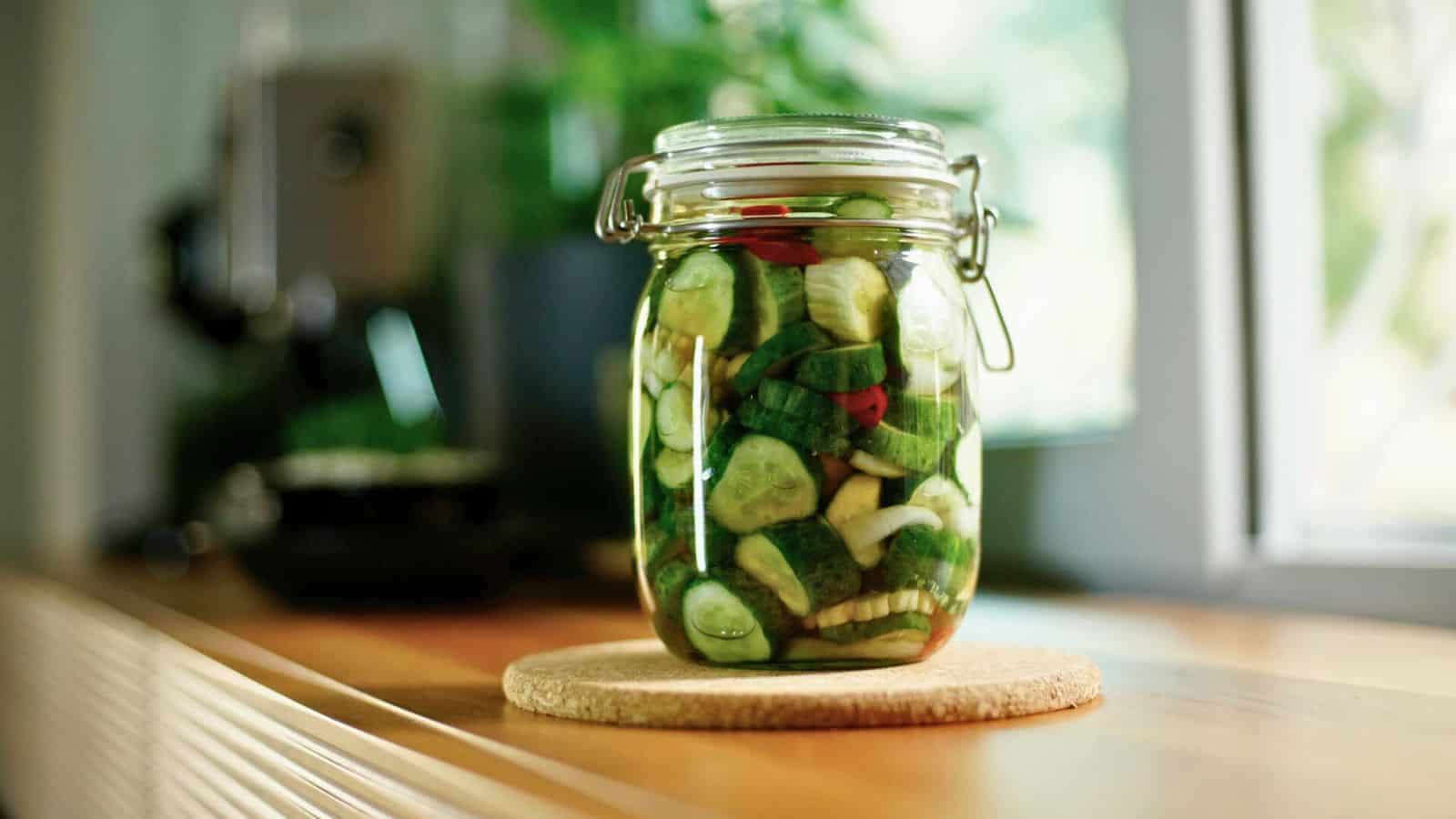 A jar of pickled vegetables sits on a circular cork pad on a wooden surface. The jar contains sliced cucumbers, peppers, and other vegetables. A blurred background with a window and greenery is visible.