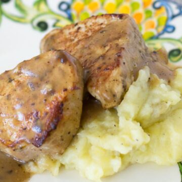 A decorative plate with a serving of mashed potatoes topped with two pieces of grilled meat smothered in brown gravy. The plate features a colorful, ornate design with green and yellow patterns around the edge—perfect for those looking to bookmark recipes that break takeout addiction.