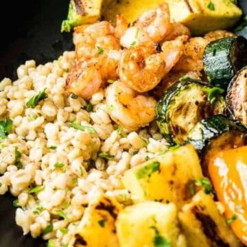 A black bowl contains a meal of grilled shrimp, zucchini slices, bell pepper pieces, and pineapple chunks, alongside a portion of cooked barley. The ingredients are garnished with green herbs. This mouthwatering dish is one to bookmark for those looking to break their takeout addiction with healthy dinner recipes.
