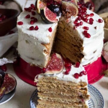 A cream-covered layer cake adorned with pomegranate seeds and figs is displayed on a table. One slice is cut and placed on a blue and white plate in front. Surrounding the cake are figs in a bowl, a white cloth, and other decorative elements.