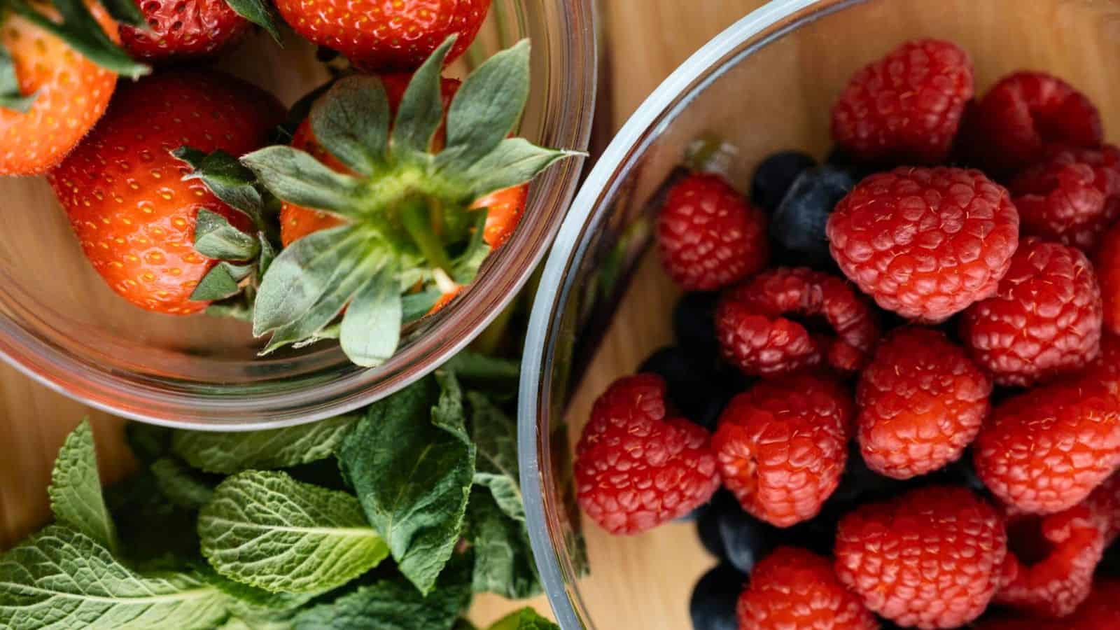 Two bowls of strawberries, blueberries, and raspberries placed on top of table with a leavy green in the background