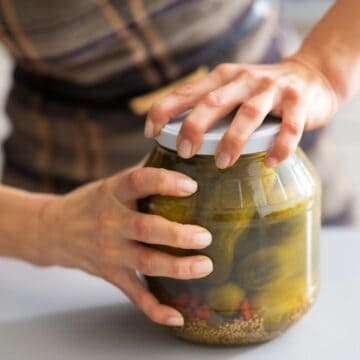 A person wearing a plaid dress is holding a large glass jar of pickles with both hands on a kitchen counter. The jar contains pickles, mustard seeds, and red chilies.