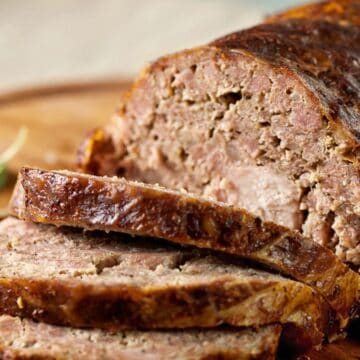 A close-up of a sliced meatloaf on a wooden cutting board, its browned crust giving way to a moist, tender interior. Some leaves for garnish are placed next to it.
