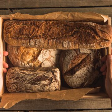 A person holds a wooden tray filled with an assortment of rustic bread loaves, including a baguette and several round and rectangular loaves, all placed on a wooden surface.
