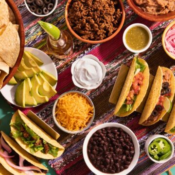 A variety of Mexican foods are arranged on a colorful tablecloth. There are hard shell tacos filled with different ingredients, bowls of black beans, shredded cheese, avocado slices, and salsa. Tortilla chips and various sauces are also present.