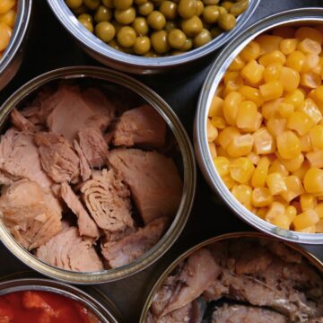 An assortment of canned goods displayed from a top-down view. The opened cans contain items such as corn, peas, tuna, beans, and other vegetables. The contents of each can are clearly visible and arranged closely together.