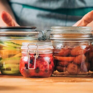 A person holding three glass jars filled with preserved foods. The jars contain sliced cucumbers, red berries, and a mixture of vegetables in liquid. The jars rest on a wooden counter with lemons, an apple, and a metal bowl nearby.