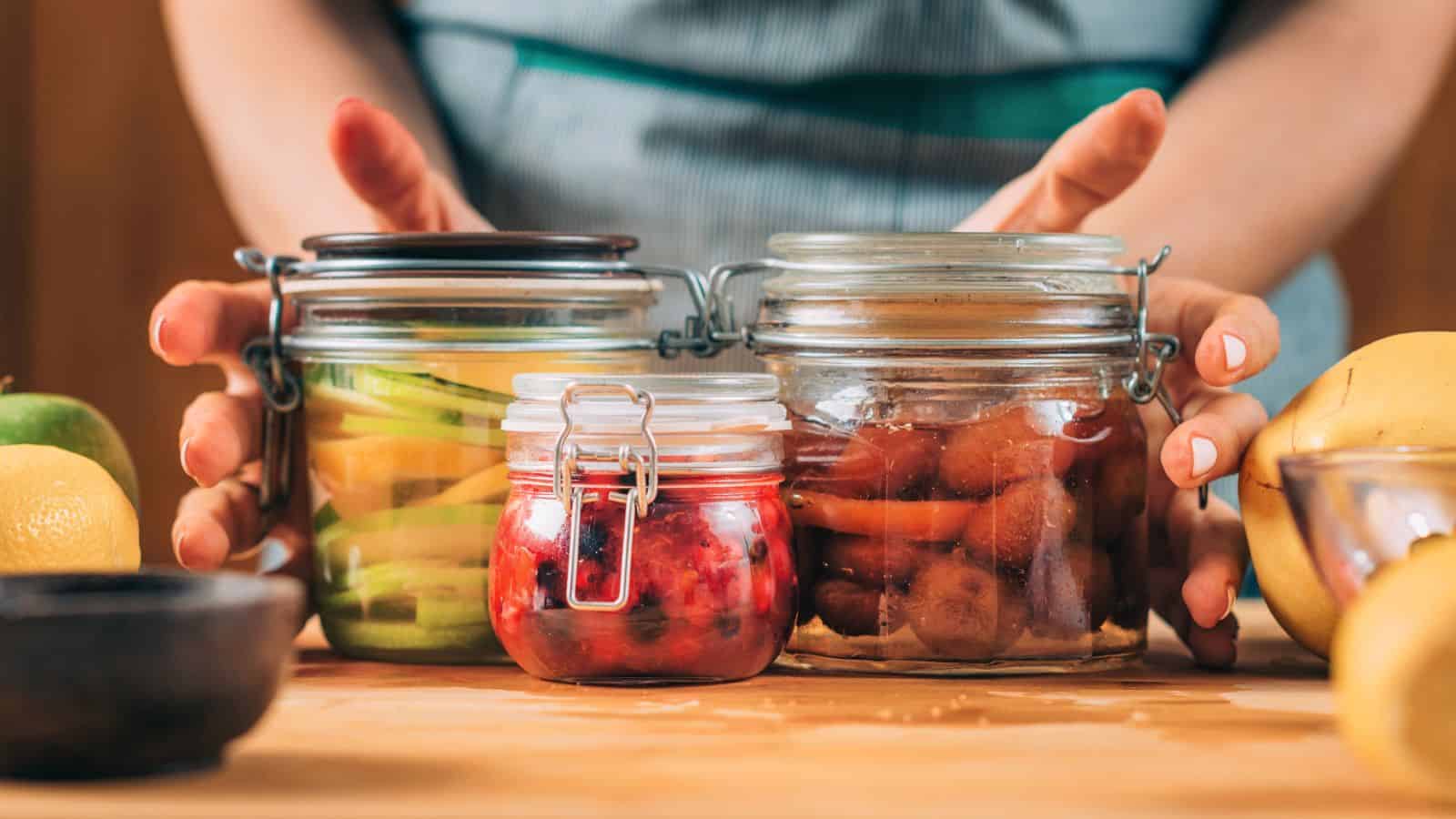 A person holding three glass jars filled with preserved foods. The jars contain sliced cucumbers, red berries, and a mixture of vegetables in liquid. The jars rest on a wooden counter with lemons, an apple, and a metal bowl nearby.
