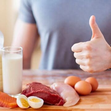 A person with a thumb up is standing behind a wooden cutting board with various foods on it. The foods include a bottle and glass of milk, raw salmon, raw beef, raw chicken, two eggs, and a sliced egg. The person is wearing a gray shirt and a fitness tracker.