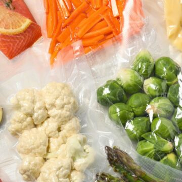 An assortment of vacuum-sealed foods is laid out including sliced eggplant, lemon with herbs, carrot sticks, cheese, cherry tomatoes, cauliflower, Brussels sprouts, asparagus, and sausages. Each item is individually wrapped in clear plastic packaging.