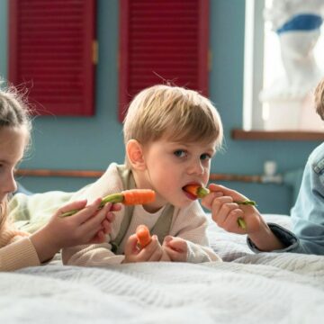 Three children are on a bed with white sheets. The young girl on the left and the boy on the right are feeding carrot sticks to the toddler in the center. The background includes red shutters and a white statue.
