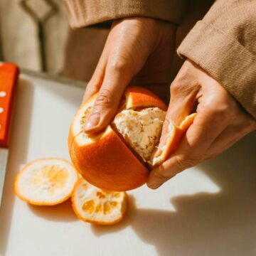 A person is peeling an orange with both hands. A knife with a red handle rests on the white cutting board next to the orange slices. The person, clad in a brown jacket, works with focus against a slightly blurred background.