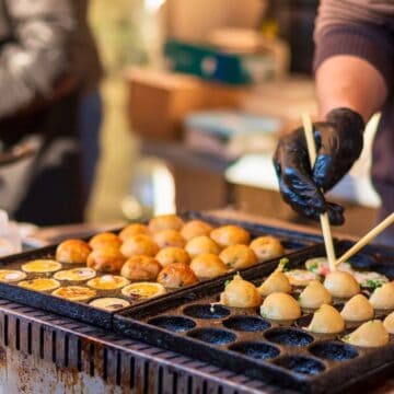 A person with chopsticks cooking golden brown takoyaki balls on a griddle at a food stall. The griddle has multiple spherical molds, and various sauces and condiments are visible in the background.