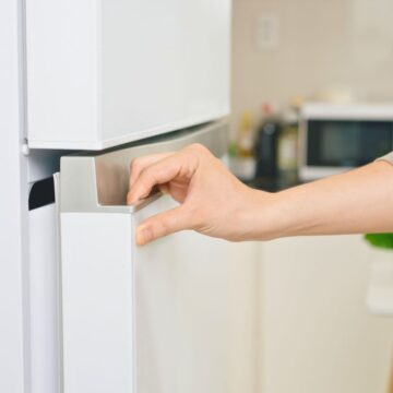 A person is opening the freezer compartment of a white refrigerator in a kitchen. The background features a kitchen counter with a microwave, various bottles, and fresh vegetables such as bell peppers and leafy greens.