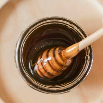 A hand holding a honey dipper is seen dipping it into a glass jar filled with dark-colored honey. The jar is placed on a wooden tray, and the background is blurred.