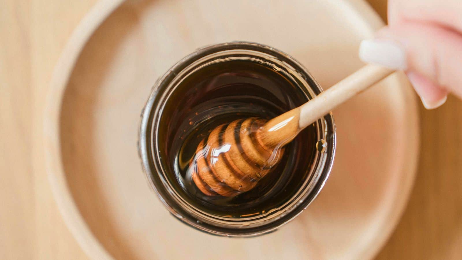 A hand holding a honey dipper is seen dipping it into a glass jar filled with dark-colored honey. The jar is placed on a wooden tray, and the background is blurred.