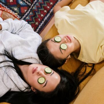 Two women lying down on carpet with cucumber slices placed over their closed eyes