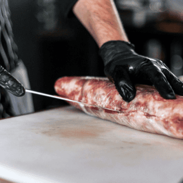 A person wearing black gloves is slicing a large piece of raw meat on a white cutting board using a knife. The person is dressed in a striped apron, and the background appears to be a kitchen or food preparation area.