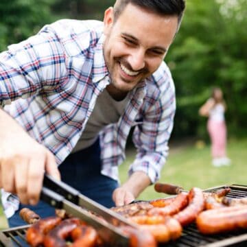 A man is grilling sausages on a barbecue grill outdoors. Wearing a plaid shirt and smiling, he uses tongs to turn the sausages with ease.