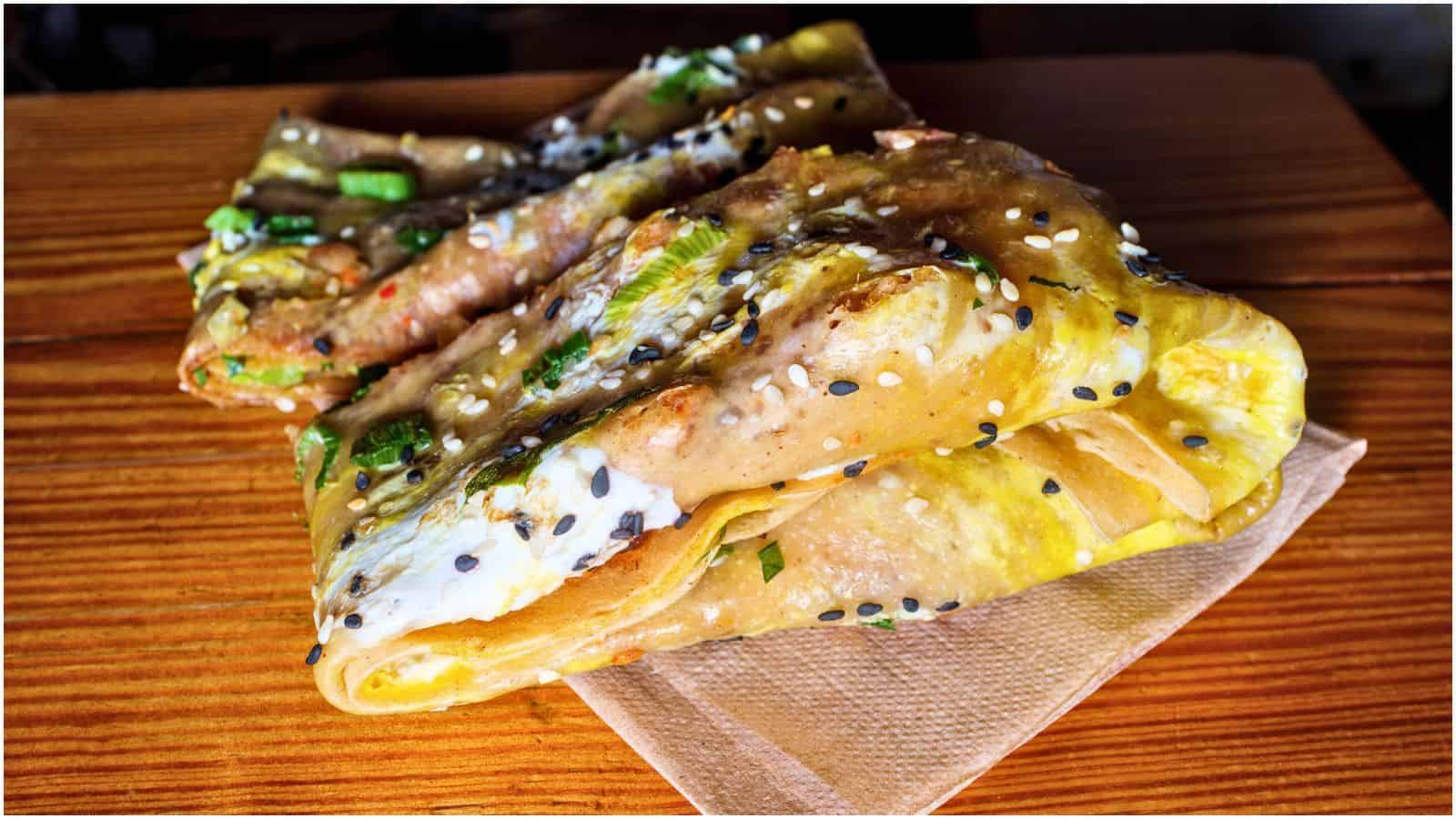 A neatly folded jianbing, one of the iconic street foods, commonly made with a crepe-like base, egg, scallions, and a variety of fillings. It is sprinkled with sesame seeds and rests on a brown napkin atop a wooden table.