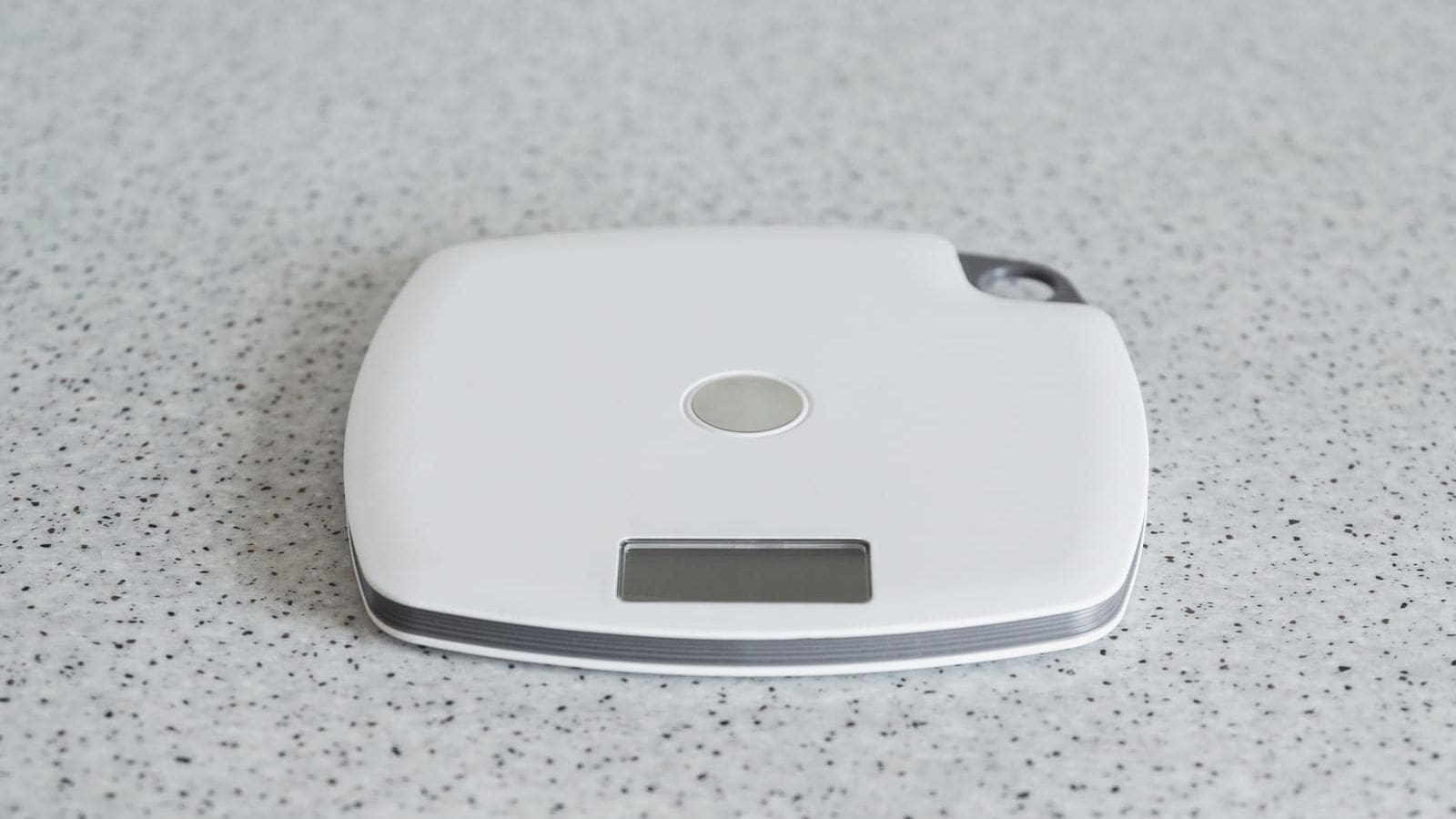 A compact white digital scale with a circular button in the center and a small rectangular LCD screen at the bottom sits on a speckled gray surface.