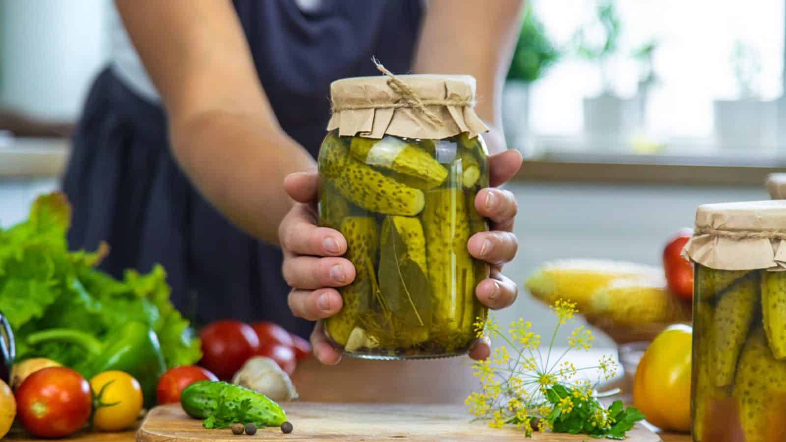 Person holding up a jar of pickled vegetables with other vegetables and a chopping board in the background