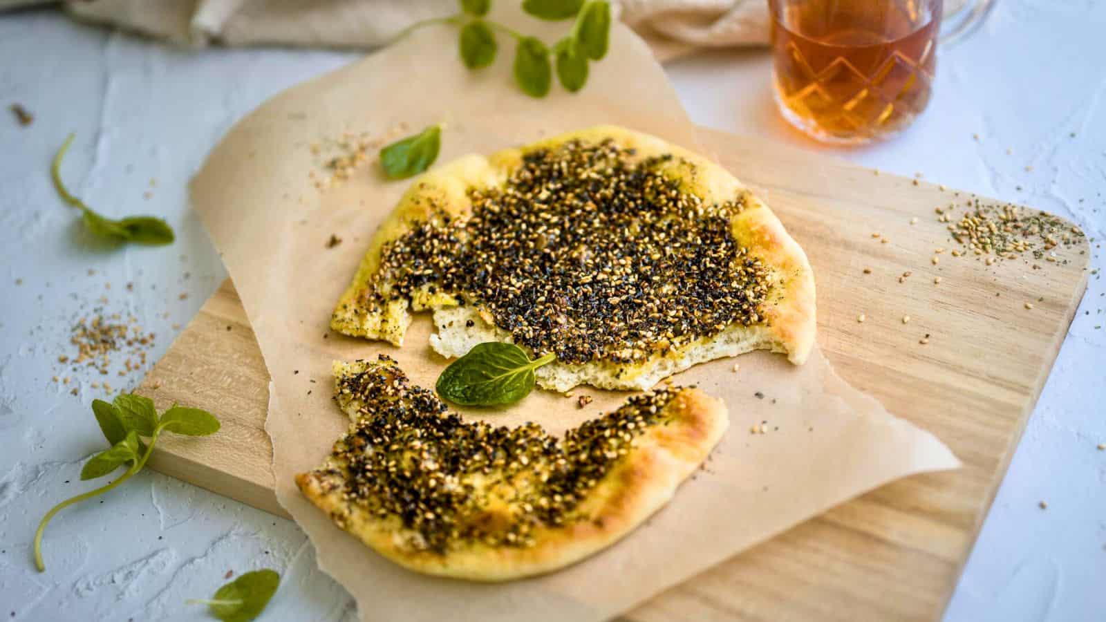 Flatbread topped with za'atar seasoning is partially cut, garnished with fresh leaves on a wooden cutting board. A napkin and a glass of tea are visible in the background. Scattered sesame seeds and mint leaves.