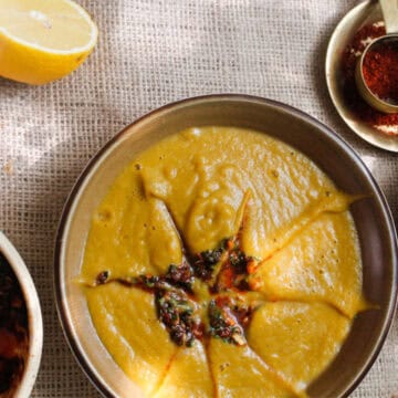 A bowl of yellow soup garnished with herbs and spices is placed on a textured fabric surface.
