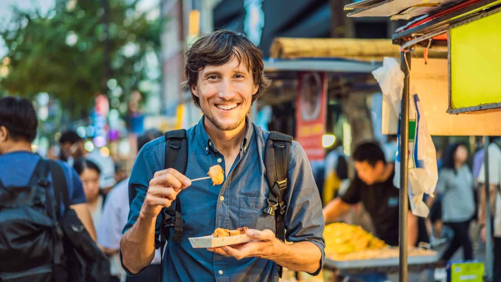 A man stands outdoors at a food market, smiling while enjoying iconic street foods from a skewer. He holds a small paper tray with additional items. Wearing a backpack and a blue shirt, the traveler is surrounded by the bustling market filled with people and food stalls in the background.