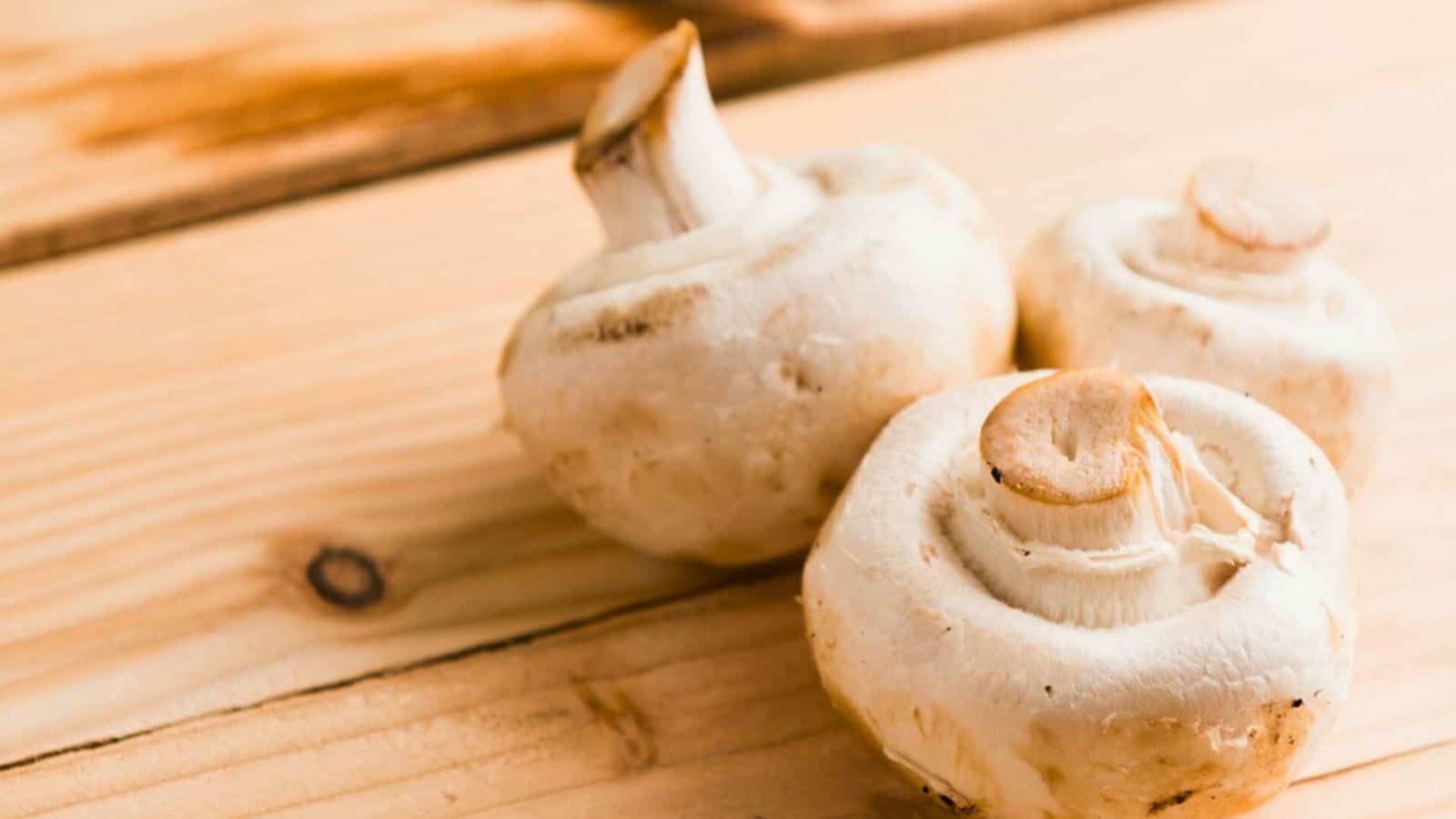 Three white mushrooms with stems attached are placed on a wooden surface, showcasing their slightly rough texture. The wooden background has visible grain patterns and knot details.
