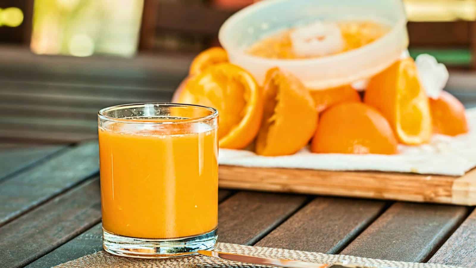 A glass of orange juice sits on a table in the foreground. In the background, there are several halved oranges placed on a wooden board.