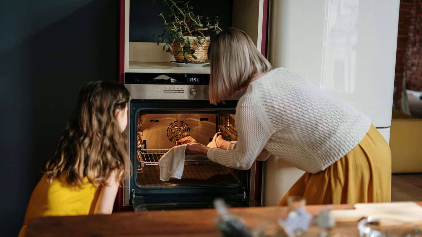 A woman and a young girl are baking together in a bright, homey kitchen. The woman, wearing a white sweater and yellow skirt, places a dish into the oven while the girl in a yellow dress watches intently. A potted plant sits atop the oven.