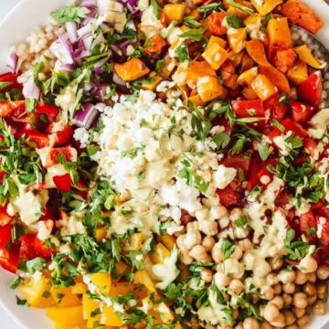 A colorful salad in a white bowl containing chopped red and yellow bell peppers, roasted sweet potatoes, chickpeas, red onions, tomatoes, fresh herbs, and a dollop of a creamy dressing, with a silver spoon resting on the side. An orange napkin is to the right.