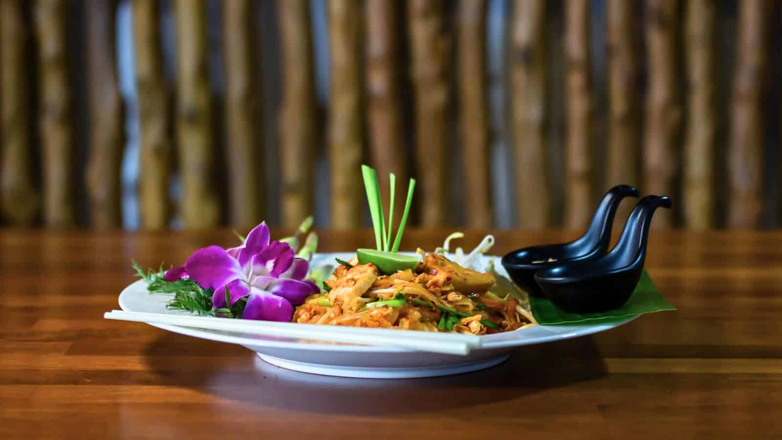 A white plate with a serving of stir-fried noodles garnished with green onions, a purple orchid, and a slice of lime sits on a wooden table. Two black sauce dishes are placed on the plate's edge, with a green leaf underneath.